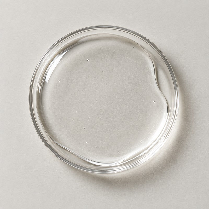 A shallow glass dish holding a pool of clear gel on a stone grey background