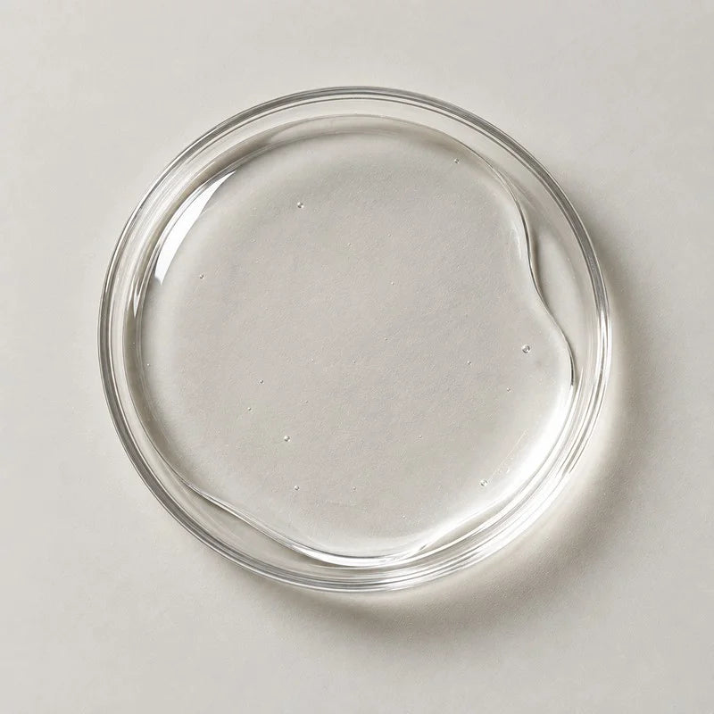 A shallow glass dish holding a pool of clear gel on a stone grey background